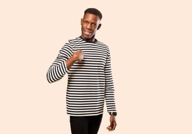 Genç Afrikalı Amerikalı siyah adam mutlu, gururlu ve şaşırmış görünüyor, neşeyle kendini işaret ediyor, kendinden emin ve bej duvara karşı yüce hissediyor.