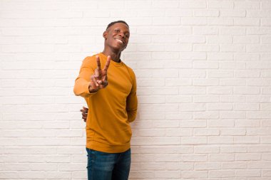 young african american black man smiling and looking happy, carefree and positive, gesturing victory or peace with one hand against brick wall