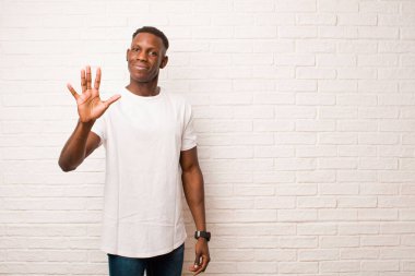 Genç Afro-Amerikan siyahi adam gülümsüyor ve dost canlısı görünüyor. 5 ya da 5 numarayı gösteriyor. Eli önde, tuğla duvara doğru sayıyor.