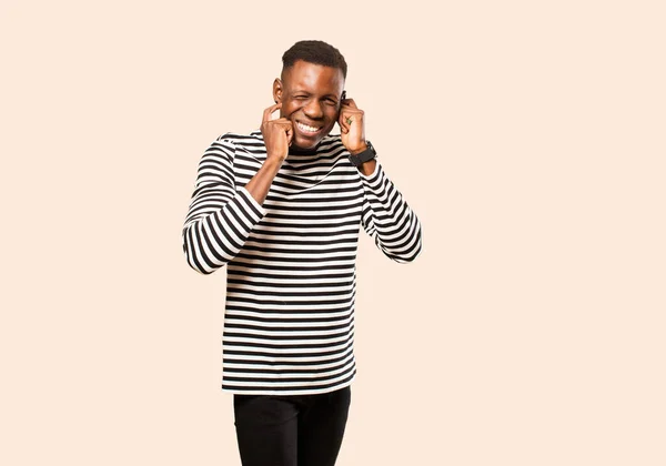 young african american black man looking angry, stressed and annoyed, covering both ears to a deafening noise, sound or loud music against beige wall