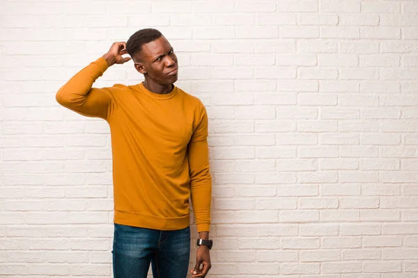 young african american black man feeling puzzled and confused, scratching head and looking to the side against brick wall