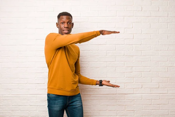 young african american black man smiling, feeling happy, positive and satisfied, holding or showing object or concept on copy space against brick wall