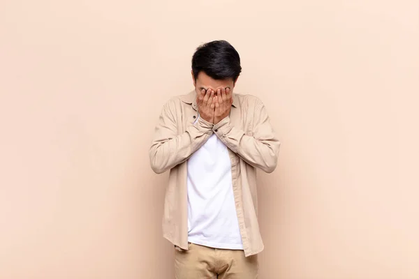 若い中国人男性は悲しみ 落ち込んで両手で顔を覆い平らな色の壁に向かって泣いています — ストック写真