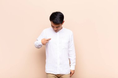 Genç Çinli adam neşeyle ve gelişigüzel gülümsüyor, aşağıya bakıyor ve düz renkli duvara doğru göğsünü işaret ediyor.