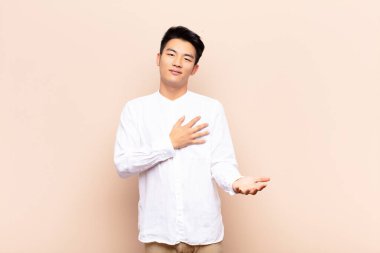 Genç Çinli adam mutlu ve aşık, bir eli kalbinin yanında gülümsüyor ve diğeri düz renkli duvara yaslanmış.