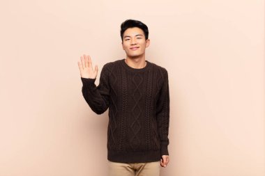 Genç Çinli adam mutlu ve neşeyle gülümsüyor, el sallıyor, seni selamlıyor, ya da düz renkli duvara veda ediyor.