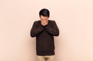 Genç Çinli adam üzgün, hayal kırıklığına uğramış, gergin ve depresif, iki eliyle yüzünü kapatıyor, düz renkli duvara karşı ağlıyor.