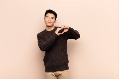 Genç Çinli adam gülümsüyor ve mutlu, sevimli, romantik ve aşık, iki elini de düz duvara dayayıp kalp şekli veriyor.