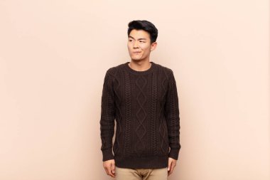 Genç Çinli adam şaşkın ve şaşkın görünüyor, merak ediyor ya da bir sorunu çözmeye çalışıyor ya da düz renkli duvara karşı düşünüyor.