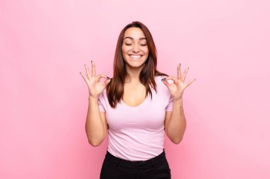 Genç ve güzel bir kadın konsantre olmuş ve meditasyon yapıyor. Tatmin olmuş ve rahatlamış hissediyor. Pembe duvara karşı bir seçim yapıyor.