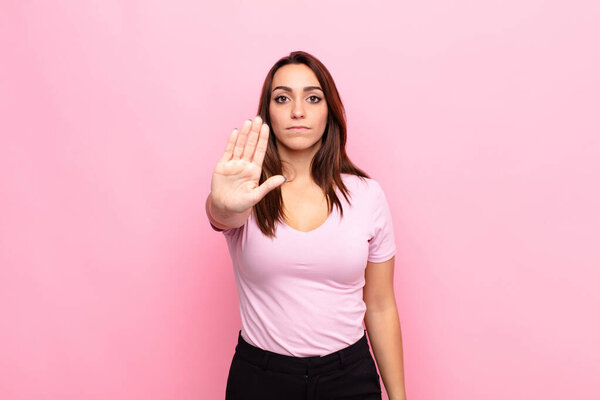 young pretty woman looking serious, stern, displeased and angry showing open palm making stop gesture against pink wall