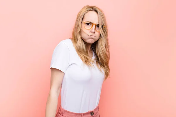 Young pretty blonde woman with a goofy, crazy, surprised expression, puffing cheeks, feeling stuffed, fat and full of food against flat color wall — Stock Image Young Pretty Blonde Woman Goofy Crazy Surprised Expression Puffing Cheeks Stock Image