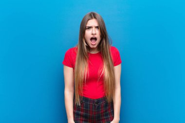 Genç ve güzel bir kadın korkmuş ve şok olmuş hissediyor, ağzı açık mavi duvara karşı şaşkınlık içinde.