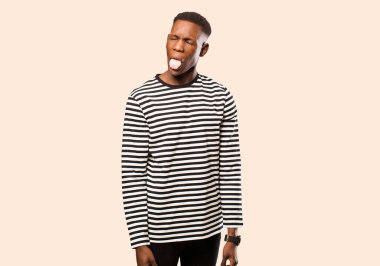 Genç Afro-Amerikan siyah adam neşeli, kaygısız, asi tavırlı, şakacı ve dilsiz, bej rengi duvarlara karşı eğleniyor.