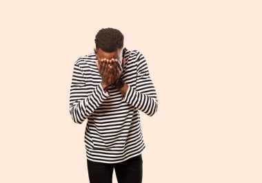 Genç Afro-Amerikan siyahi adam üzgün, hayal kırıklığına uğramış, gergin ve depresif hissediyor, yüzünü iki eliyle kapatıyor, bej rengi duvara karşı ağlıyor.