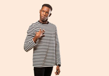 Genç Afrikalı Amerikalı siyah adam neşeyle gülümsüyor, mutlu, tatmin olmuş ve rahatlamış, kollarını kavuşturup bej rengi duvara bakıyor.