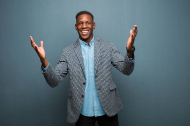 Genç Afrikalı Amerikalı siyahi adam mutlu, şaşkın, şanslı ve şaşırmış hissediyor. İki eli havada, grunge duvarına karşı zaferi kutluyor.