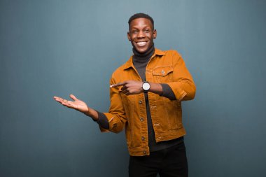 Genç Afro-Amerikan siyahi adam neşeyle gülümsüyor ve yan taraftaki fotokopi alanını işaret ediyor, grunge duvarına karşı bir nesne gösteriyor veya reklamını yapıyor.