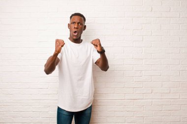 Genç Afrikalı Amerikalı siyahi adam inanılmaz bir başarıyı kutluyor. Kazanan gibi, heyecanlı ve mutlu görünüyor. Al bakalım! tuğla duvara karşı