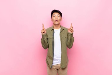 Genç Çinli adam şaşırmış, şaşırmış ve ağzı açık görünüyor, iki eliyle yukarıya bakıyor düz renkli duvara uzayı kopyalamak için.