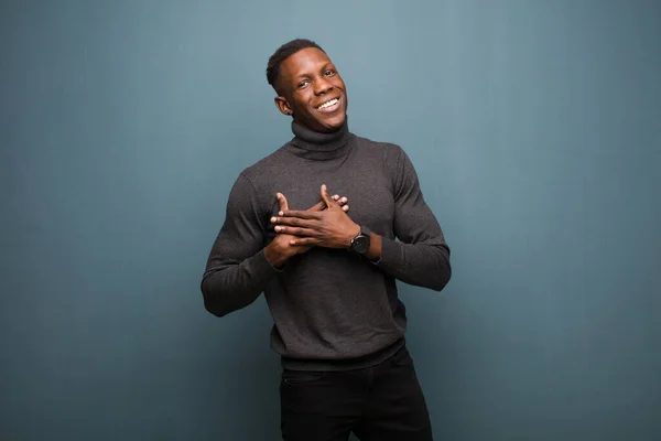 young african american black man feeling romantic, happy and in love, smiling cheerfully and holding hands close to heart against grunge wall