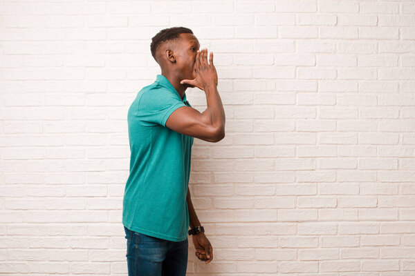 Young african american black man yelling loudly and angrily to copy space on the side, with hand next to mouth against brick wall