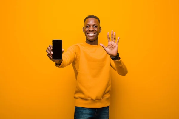 若いアフリカ系アメリカ人の黒人男性がオレンジの壁にスマートフォンを向けて — ストック写真