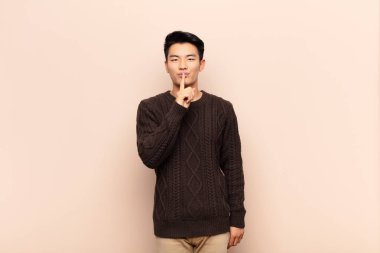 Genç Çinli adam ciddi görünüyor ve parmaklarını dudaklarına bastırıp sessizlik ya da sessizlik talep ediyor. Düz renkli duvara karşı bir sır saklıyor.