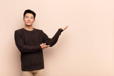 Genç Çinli adam gururla ve kendinden emin bir şekilde gülümsüyor, mutlu ve tatmin olmuş hissediyor ve düz renkli duvara karşı fotokopi alanında bir konsept gösteriyor.