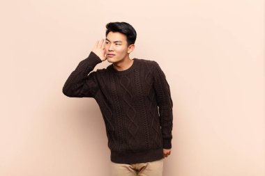 Genç Çinli bir adam tuhaf bir şekilde gülümsüyor, yan tarafa bakıyor, dedikoduları dinlemeye çalışıyor ya da düz renkli duvarlara karşı bir sır duymaya çalışıyor.