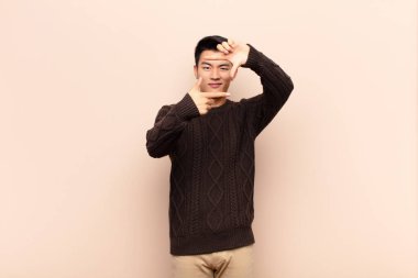 Genç Çinli adam mutlu, arkadaş canlısı ve pozitif hissediyor, gülümsüyor ve düz renkli duvara karşı elleriyle bir portre veya fotoğraf çerçevesi yapıyor.