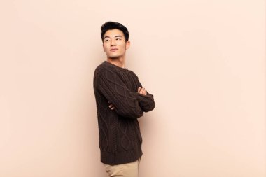 Genç Çinli adam neşeyle gülümsüyor, mutlu, tatmin olmuş ve rahatlamış, kollarını kavuşturup düz duvara bakıyor.