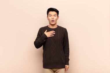 Genç Çinli adam şok olmuş, şaşırmış ve şaşırmış hissediyor, elleri göğsünde ve ağzı açık, kim diyor, ben mi? yassı renk duvarına karşı