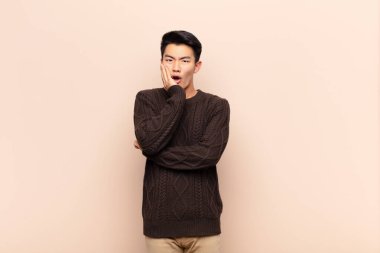 Genç Çinli adam şok ve inançsızlık içinde, elleri yanak ve kolları çapraz, sersemlemiş ve düz renkli duvara karşı şaşkın ve şaşkın hissediyor