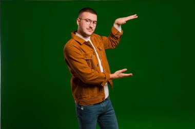 Genç yakışıklı bir adam iki eli de yan tarafında fotokopi alanı olan bir nesneyi tutuyor, düz arkaplana karşı bir nesneyi gösteriyor, sunuyor ya da reklamını yapıyor.