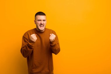 Genç yakışıklı adam sinirli, sinirli, sinirli bakışlar ve sıkı yumruklarla bağırıyor, düz arkaplana karşı öfkeli hissediyor.