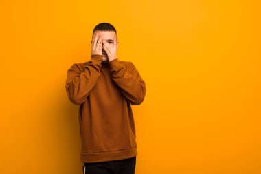 Genç yakışıklı adam elleriyle yüzünü kapatıyor, parmaklarının arasından şaşkın bir ifadeyle bakıyor ve düz arkaplana bakıyor.