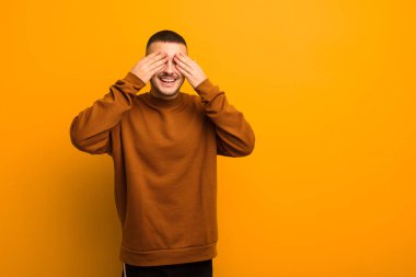 Genç yakışıklı adam gülümsüyor ve mutlu hissediyor, iki eliyle gözlerini kapatıyor ve düz arkaplanda inanılmaz bir sürpriz bekliyor.