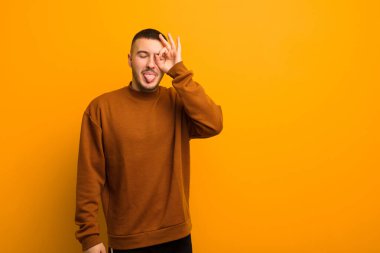 Genç yakışıklı adam mutlu bir şekilde gülümsüyor, şaka yapıyor ve gözetleme deliğinden bakıyor, düz arkaplana karşı sırları gözetliyor.