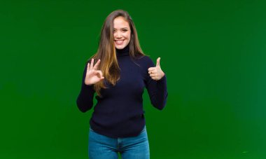 Genç, güzel bir kadın kendini mutlu, şaşkın, tatmin olmuş ve şaşırmış hissediyor. Yeşil arka planda gülümseyerek iyi ve güzel hareketler gösteriyor.