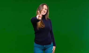 Genç ve güzel bir kadın gülümsüyor ve mutlu görünüyor, kaygısız ve pozitif, yeşil arka planda tek elle zafer ya da barış işareti yapıyor.