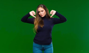 Genç, güzel bir kadın üzgün, hayal kırıklığına uğramış ya da kızgın görünüyor. Anlaşmazlıkta baş parmaklarını gösteriyor. Yeşil arka planda hayal kırıklığına uğramış hissediyor.