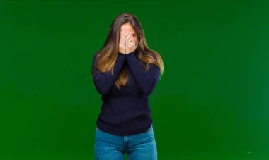 Genç, güzel bir kadın üzgün, hayal kırıklığına uğramış, gergin ve depresif, iki eliyle yüzünü kapatıyor, yeşil arka planda ağlıyor.