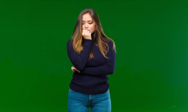 Genç ve güzel bir kadın ciddi, düşünceli ve endişeli, elleri çenesine değmiş yeşil arka plana bakıyor.