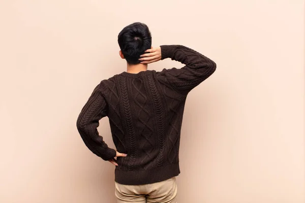 young chinese man thinking or doubting, scratching head, feeling puzzled and confused, back or rear view against flat color wall