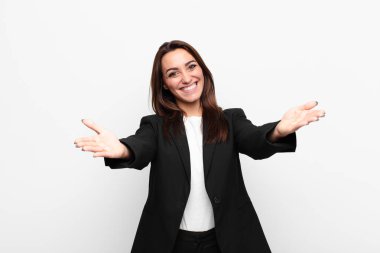 Genç, güzel iş kadını neşeyle gülümsüyor. Sıcak, dostane, sevgi dolu bir kucaklama yapıyor. Beyaz duvara karşı mutlu ve sevimli hissediyor.