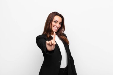 Genç ve güzel iş kadını gururla gülümsüyor ve gururla bir numaralı pozu veriyor. Beyaz duvara karşı bir lider gibi hissediyor.