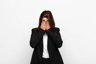 Genç, güzel iş kadını üzgün, hayal kırıklığına uğramış, gergin ve depresif hissediyor, yüzünü iki eliyle kapatıyor, beyaz duvara karşı ağlıyor.