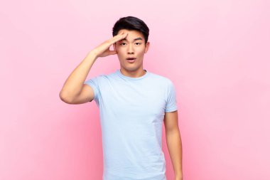 Genç Çinli adam mutlu, şaşkın ve şaşırmış görünüyor, gülümsüyor ve düz renkli duvara karşı inanılmaz ve inanılmaz iyi haberler veriyor.