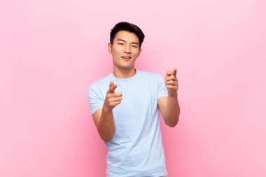 Genç Çinli adam pozitif, başarılı, mutlu bir tavırla kamerayı işaret ederek gülümsüyor, düz renkli duvara karşı elleri ile silah işareti yapıyor.